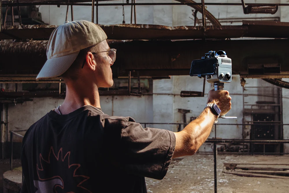 Ein Mitarbeiter von Hausgecheckt, der eine nach hinten gerichtete Kappe, eine Sonnenbrille und ein T-Shirt trägt, hält eine Kamera auf einem Stabilisator und filmt oder fotografiert in einem verlassenen Industriegebäude.